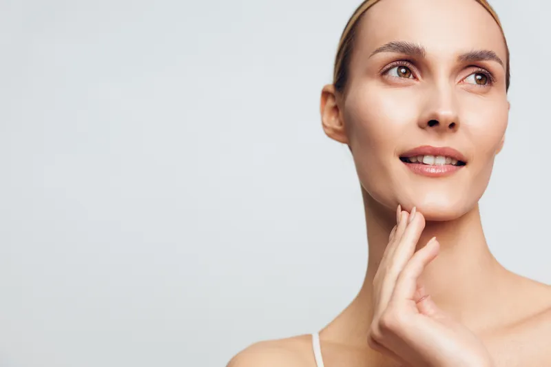 Young woman with clear skin and natural makeup touching her face gently. Soft lighting and neutral background emphasize skincare and beauty concepts.