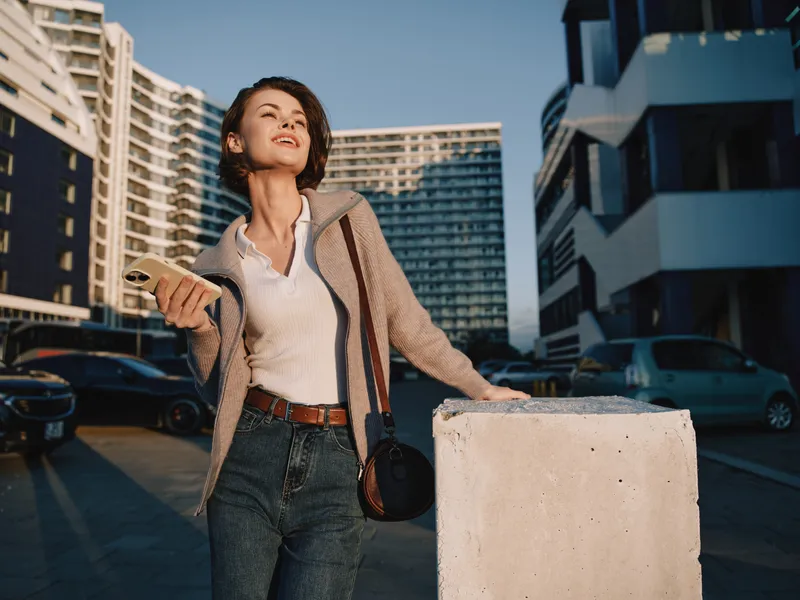 A stylish professional woman stands outdoors in a modern urban setting, smiling while texting on a smartphone. She exudes confidence, energy, and connectivity, creating a positive and dynamic