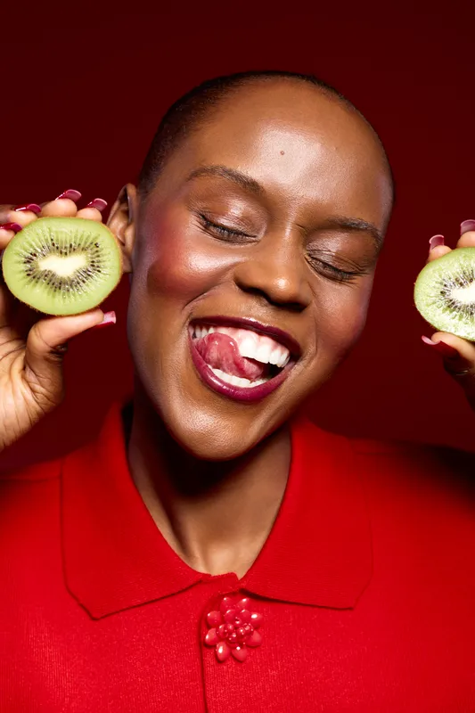 Happy African woman with dark skin holds two halves of kiwi fruit near face, smiling with eyes closed and tongue out. Vibrant red background and clothing create joyful mood and freshness concept.