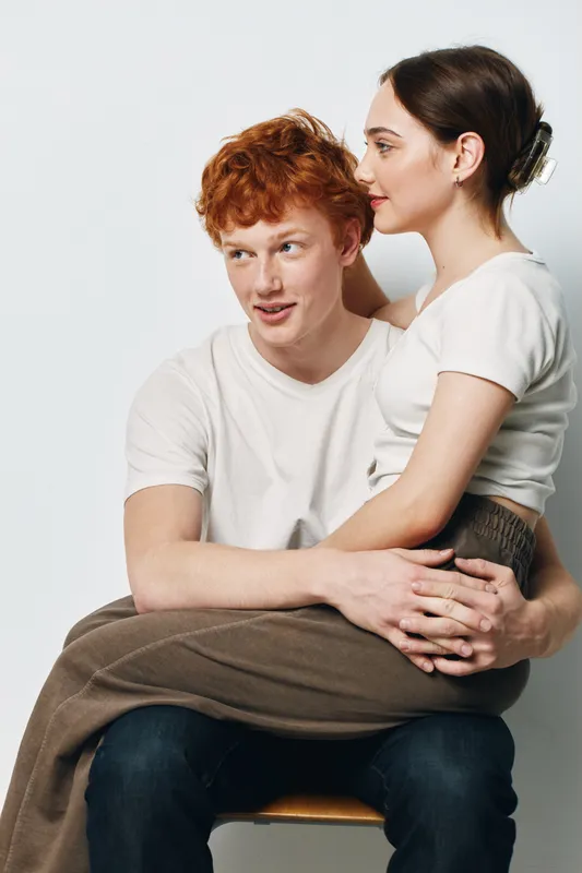 Young couple sitting with woman on man’s lap in casual clothes looking relaxed and happy with affection and connection in studio interior against white background.