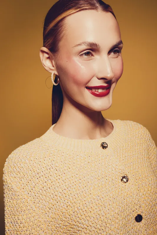 Smiling woman with red lipstick and hoop earrings wears beige knitted sweater against yellow background. Portrait of confident female model with natural makeup and sleek hairstyle.