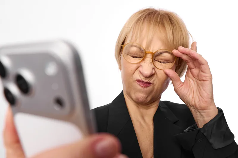 Blond woman with glasses holding a smartphone to her face, thinking and listening during a video call, office look, white background, soft studio light calm mood here