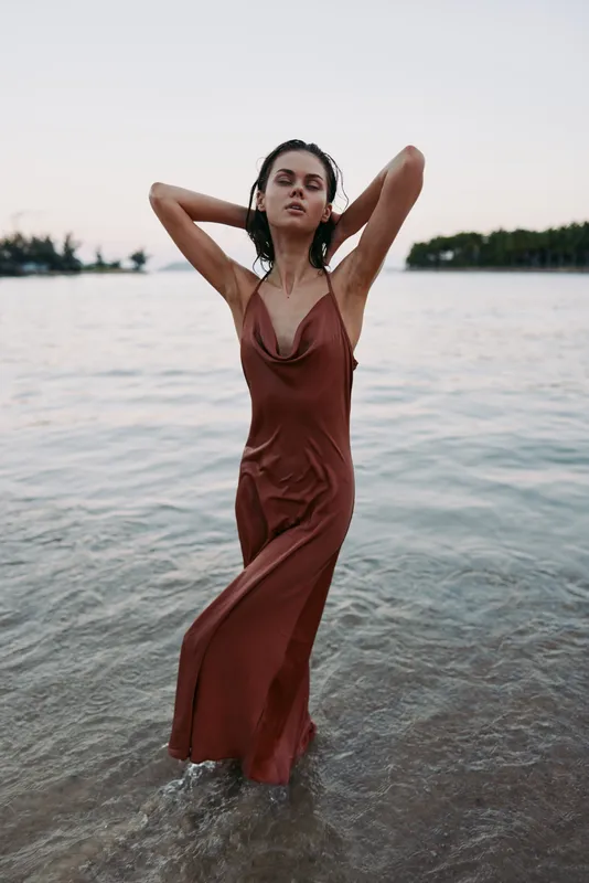 A woman in a brown slip dress stands waist-deep in calm shoreline water, striking a confident pose with hands at her head, creating a bold, natural portrait.