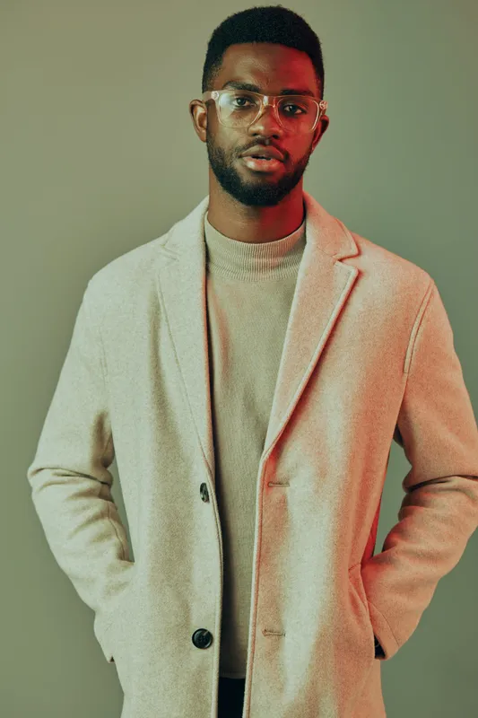 Stylish young man in a light beige coat and glasses against a muted green background, embodying modern fashion and confidence