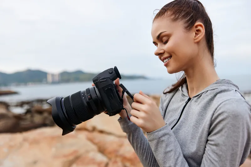 Young woman with a camera enjoying photography on the beach, dressed in a casual gray hoodie, capturing memories with a joyful smile against a serene coastal background