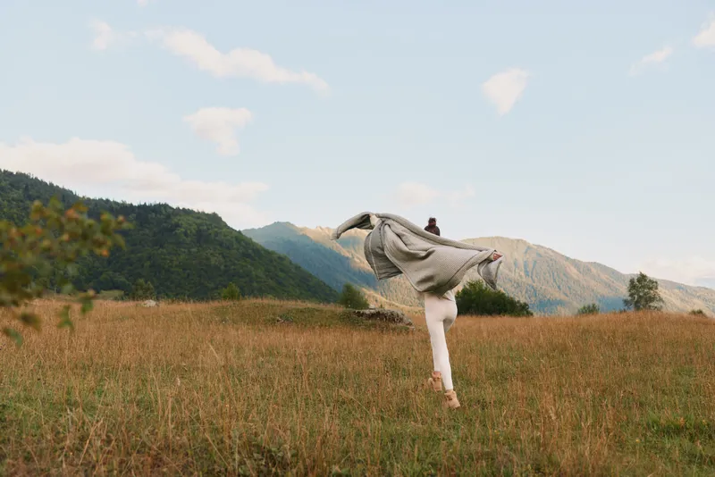 Cheerful woman outdoors, flowing scarf, joyful leap in a sunlit meadow, distant mountains, light breeze, natural colors, relaxed pose, vibrant countryside, candid moment