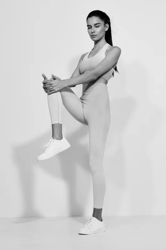 Woman sport fitness performing a standing knee stretch in monochrome, wearing athletic wear and sneakers against a plain background, showcasing strength and flexibility.