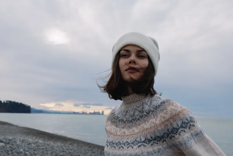 Woman wearing a knit sweater and beanie by the seaside, wind in hair, calm water and cloudy sky, casual winter fashion portrait with outdoor coastal mood