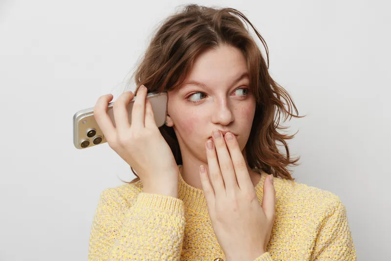 Young woman in yellow sweater holds smartphone near ear and covers mouth with hand, looking surprised. Concept for secret, gossip, communication, and curiosity.