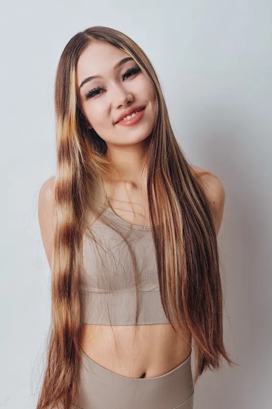 Young woman with long straight hair wearing beige top smiling gently, studio portrait with soft lighting and plain background for beauty and fashion concepts.