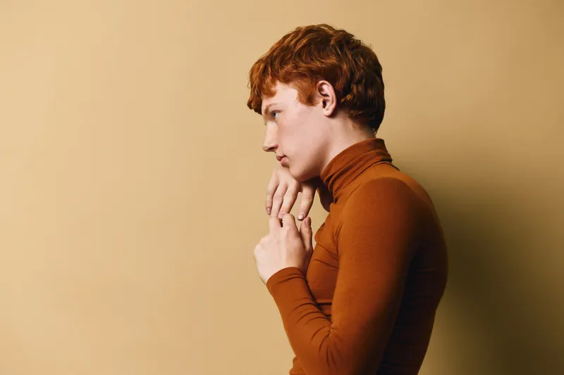 Young man with red hair wearing brown turtleneck sweater standing against beige background in profile and looking ahead with thoughtful expression in studio portrait.