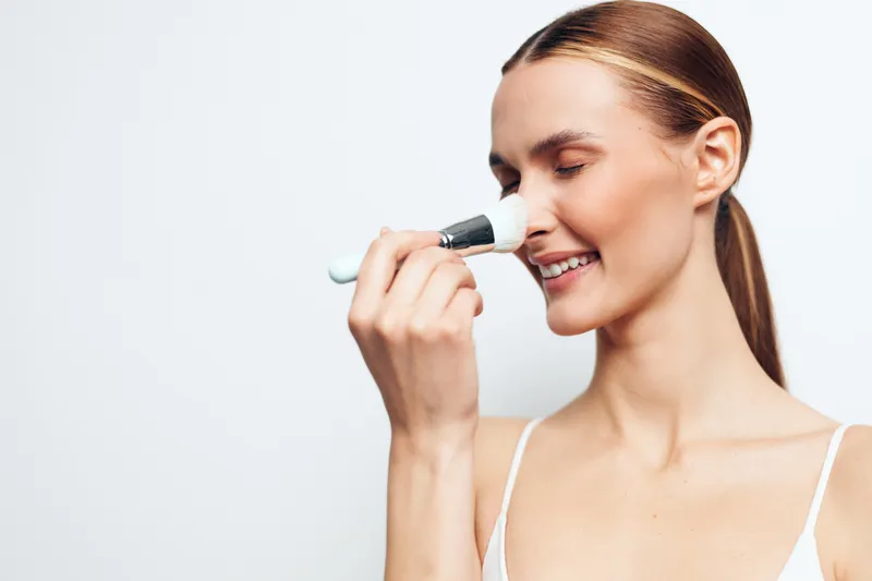 Smiling woman with clear skin applying makeup brush on her face. Close-up with white background. Concept of beauty routine, skincare, and natural look with soft lighting.