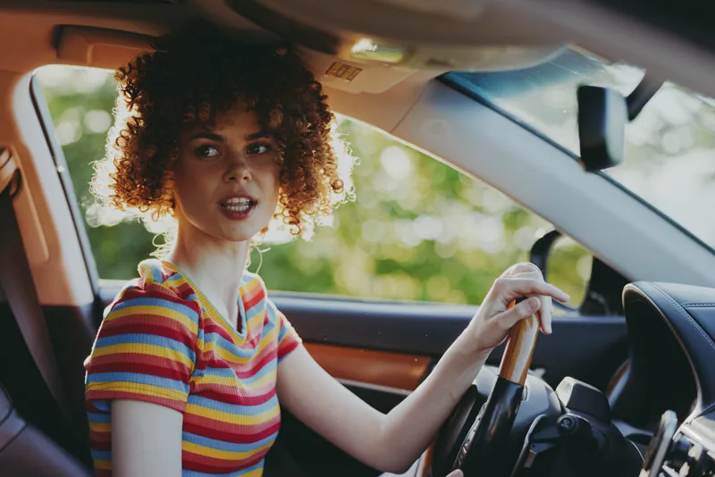 Vibrant woman driving a car, striped shirt, candid lifestyle moment, sunlit interior, retro film color mood, relaxed expression, natural tones, greenery outside, joyful motion