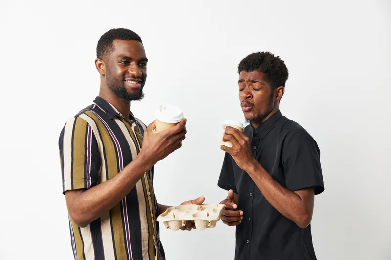 Two smiling African American men enjoy coffee together, sharing a joyful moment while dressed in casual attire against a neutral background. Perfect for lifestyle themes.