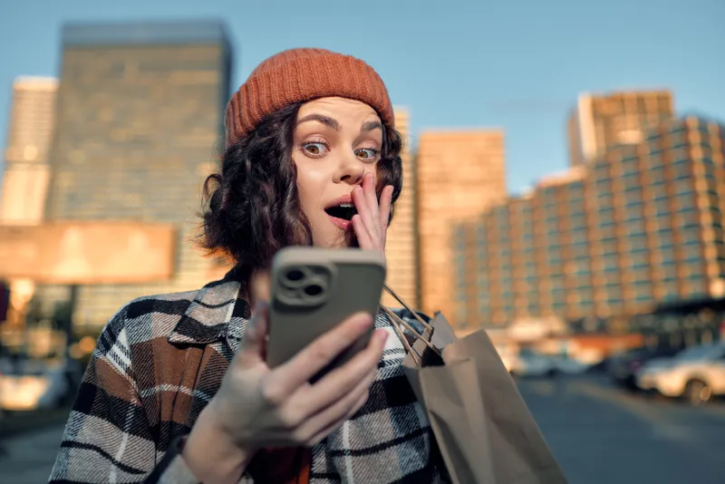 Woman with smartphone shows surprised expression while shopping in an urban portrait, candid moment emphasizing authenticity, golden hour glow, mindful living and emotional storytelling.