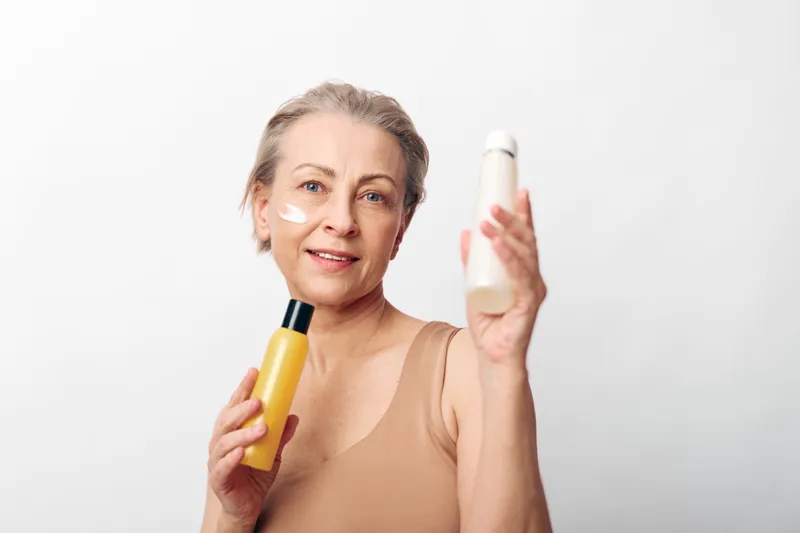 Mature woman with short hair holds two skincare bottles applying cream on cheek. Studio shot on white background. Concept of beauty care, anti aging, and natural cosmetics.