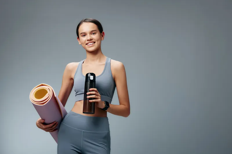 Fitness woman in athletic wear holding a water bottle and yoga mat against a neutral background, ready for a workout and wellness routine.