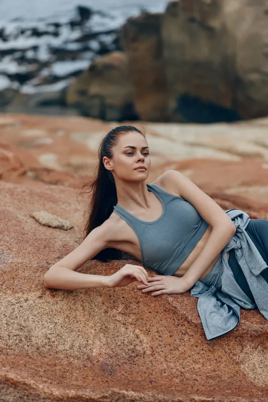 Young athletic model in a grey sports top posing on rocky beach during sunset, showcasing fitness and serenity with a natural background