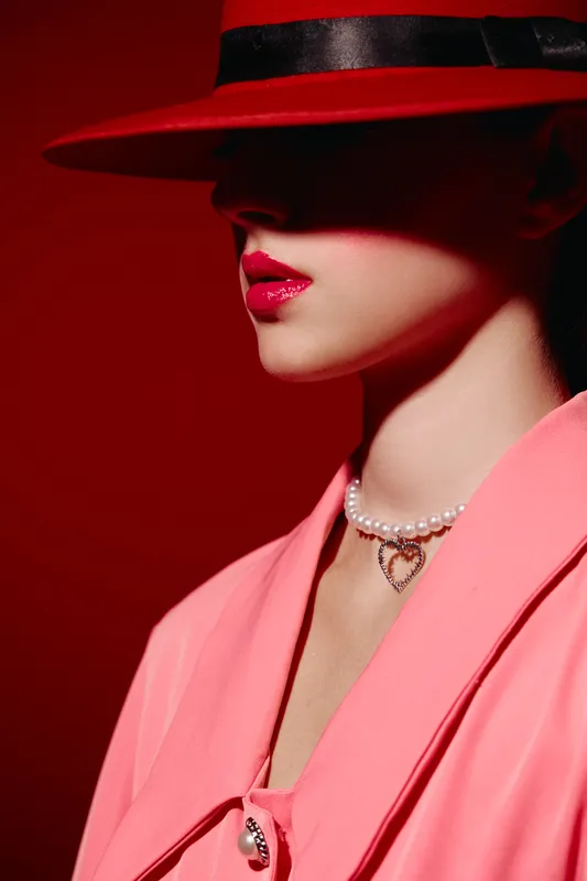 Fashionable young woman wearing a red hat that shadows her face, bright pink lipstick, pearl necklace with pendant, and coral jacket. Bold makeup complements creative beauty theme.