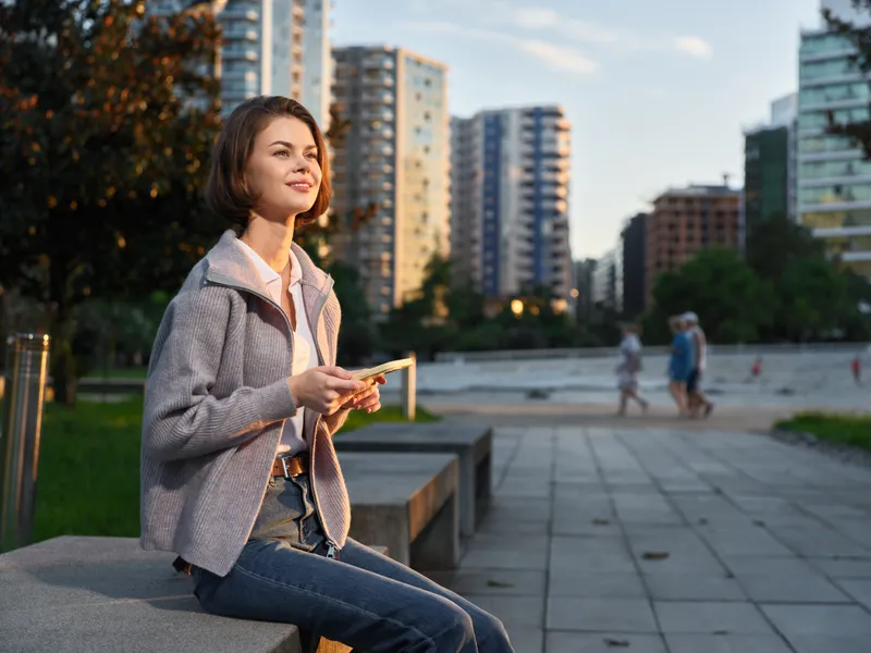A cheerful professional woman sits on a concrete ledge in a modern city park, texting on her phone with a warm smile, conveying a productive, upbeat urban vibe.