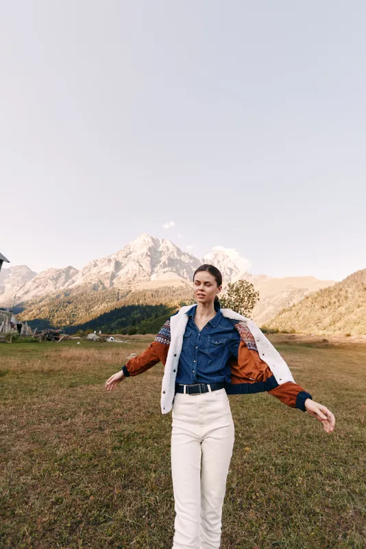 Woman in denim and jacket stands in meadow with mountains behind, portrait of travel and outdoors lifestyle, fashion model in casual outfit enjoying alpine scenery and sunlight.