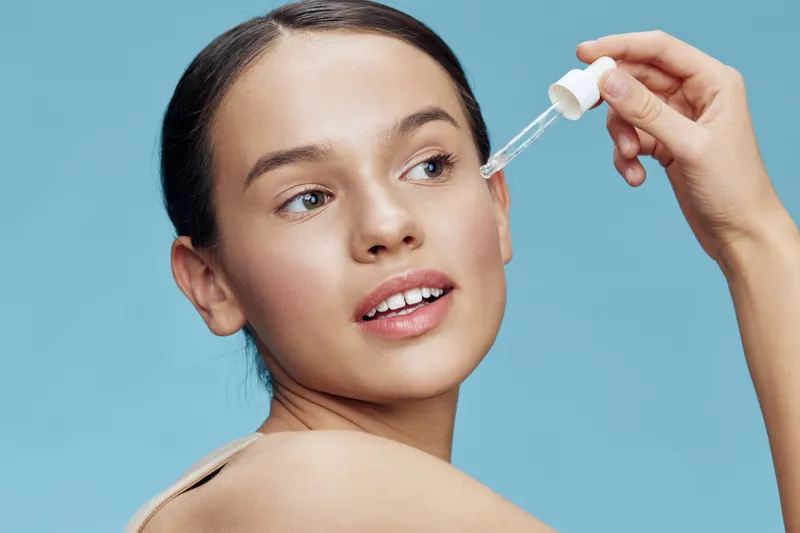 Young woman applying serum to her face, showcasing clear skin and a radiant smile against a soft blue background, emphasizing skincare and beauty