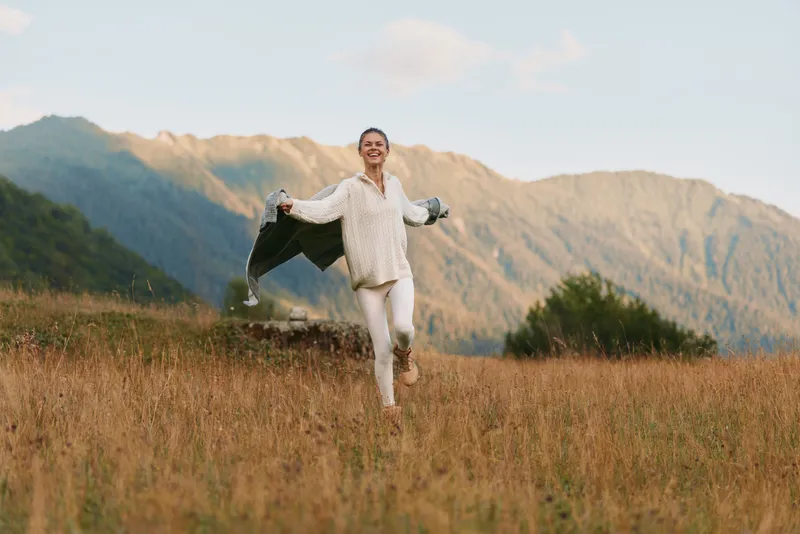 Smiling woman in a white blouse dances in a golden field with mountains behind, portraying a carefree outdoor lifestyle, joy, and freedom under bright light and glow.