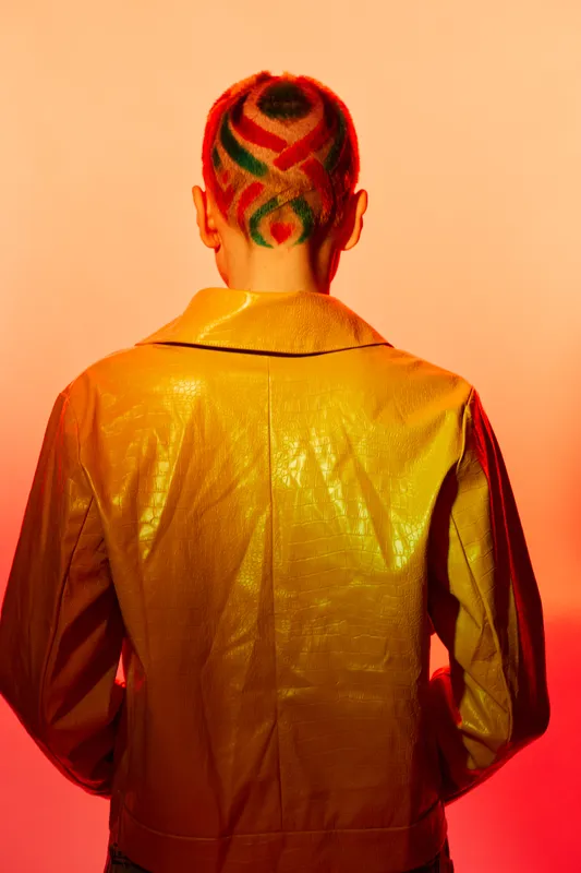 Woman with colorful shaved hairstyle seen from behind, wearing a shiny yellow jacket against an orange gradient background in stylish lighting.