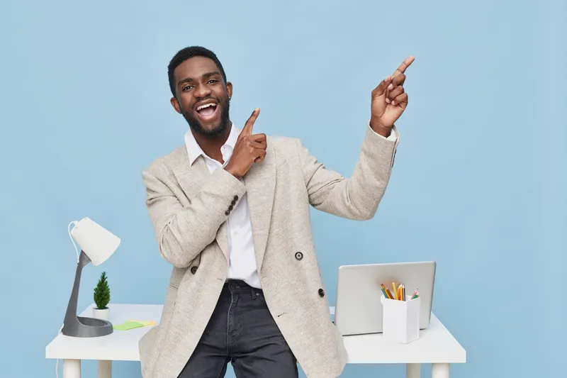 Smiling man with dark skin in casual beige sweater and white shirt pointing upwards while standing in bright modern office with white desk and laptop, cheerful expression. People lifestyle concept
