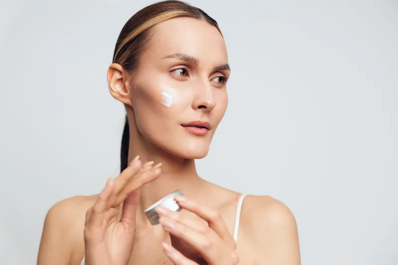 Young woman applies face cream on cheek holds jar with skincare product looks away with calm expression isolated on white background beauty routine concept.
