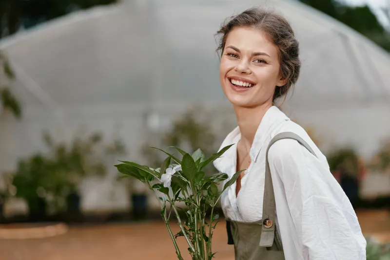 Young woman smiling holding plants in a greenhouse, showcasing vibrant greenery and a warm, inviting atmosphere, ideal for promoting gardening and sustainability