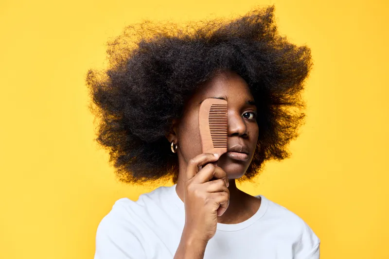 Portrait of a young African woman holding a comb in front of her eye, showcasing natural hair against a vibrant yellow background Empowerment and hair care concepts
