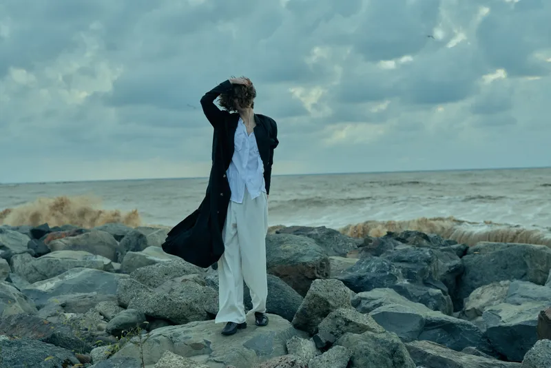 Windy sea on a rocky shore with a solitary figure in a long coat gazing to the horizon, capturing a mood of travel and introspection against stormy skies