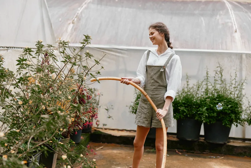 Joyful young woman watering plants in a greenhouse wearing casual overalls and a white shirt, surrounded by lush greenery, showcasing a connection to nature