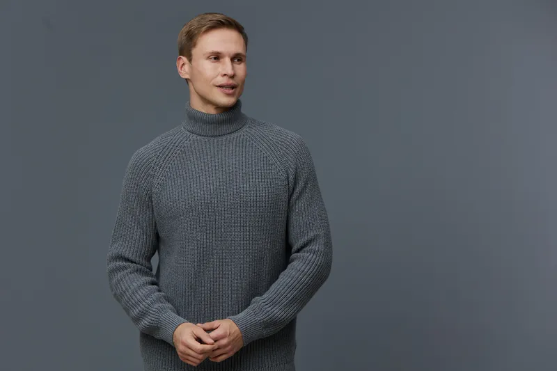 Smiling man wearing gray knitted sweater looking to the side against plain gray background, studio portrait, casual fashion, people lifestyle concept, warm clothing, confident expression, indoor setti