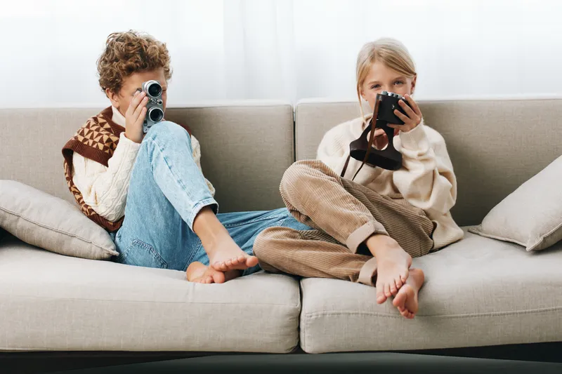 Two children sit on a sofa, each focused on a camera, creating a warm, playful moment of curiosity, exploration, and imaginative photography at home.