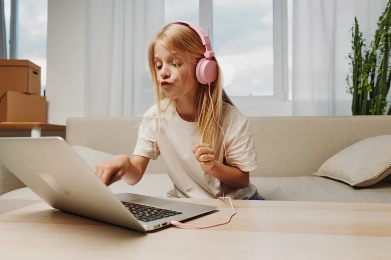 Young girl with long hair wearing pink headphones enjoys music and uses laptop at home, sitting on sofa in bright living room.
