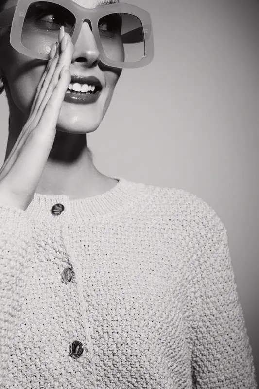 Smiling white woman with fair skin wearing oversized glasses and knitted sweater holds hand near mouth in black and white studio portrait. Concept for fashion and communication.