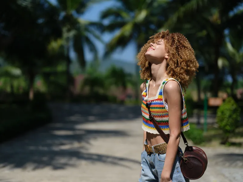 young woman with curly hair wearing colorful top and denim shorts standing outdoors, enjoying warm sunlight with eyes closed, tropical palm trees in background