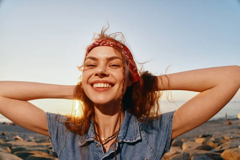 smiling woman with curly hair wearing a red headband and denim shirt enjoying sunset on rocky beach with golden light