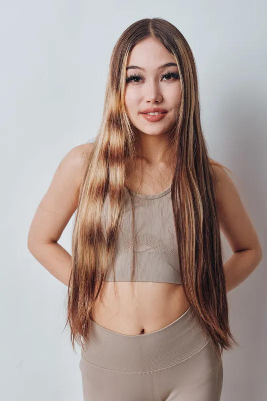 Young woman with long straight hair wearing beige sleeveless top and pants posing with hands behind back against plain white background in studio.