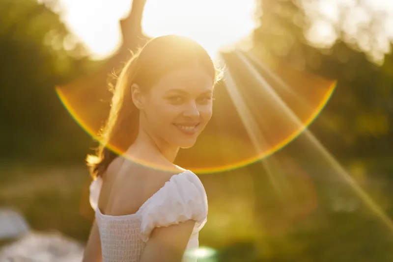 Young woman smiling in a sunlit outdoor setting, wearing a white dress, exuding joy and warmth with a vibrant bokeh background