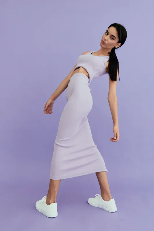 young woman in lilac matching outfit poses uniquely against purple background, showcasing casual style with white sneakers and long black hair