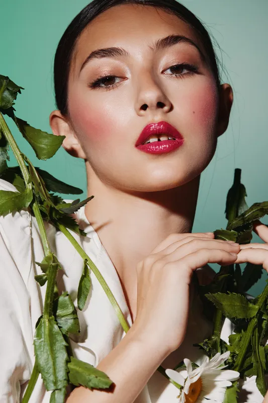 Young female model with vibrant makeup posing confidently with dramatic lighting while holding green plant leaves. Creative beauty and fashion concept with glossy lips and artistic styling.