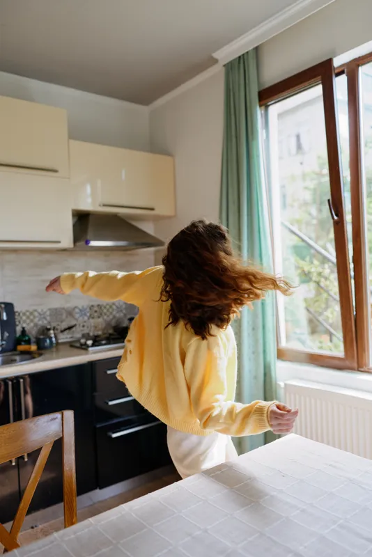 A young woman in a yellow sweater dances in a bright kitchen, her back to the camera, conveying carefree movement and solo joy in a cozy daily moment.