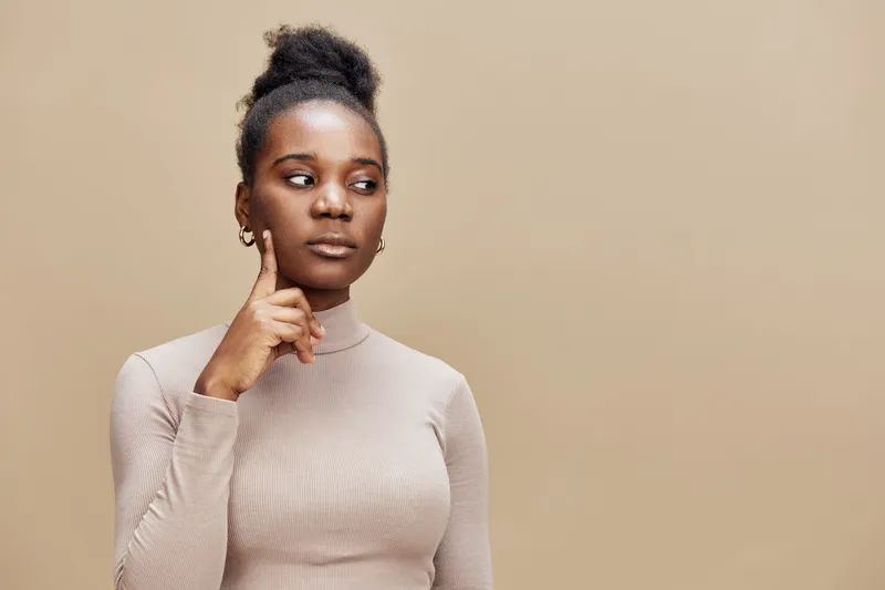 Thoughtful young Black woman in beige outfit with neutral background, expressing curiosity and contemplation through her pose and expression, perfect for lifestyle concepts