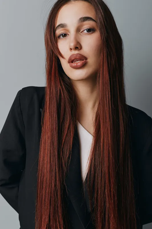 Woman with long straight hair and natural makeup posing in black blazer against gray background. Fashion portrait with serious expression and smooth skin closeup in studio lighting.