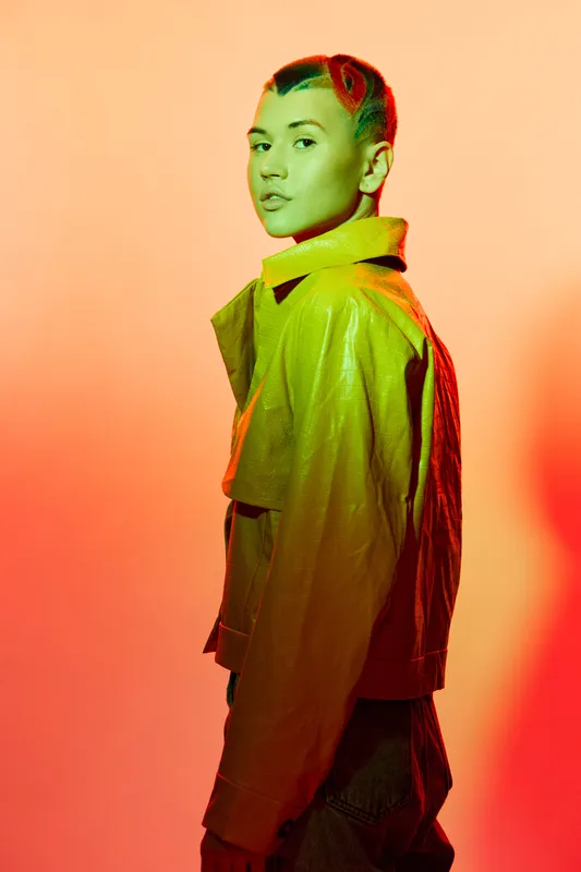 Young woman with creative hairstyle wearing yellow jacket poses under colorful lighting, expressing modern style and bold fashion in studio setting.