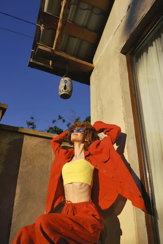 A stylish young woman wears a bright yellow crop top and red outfit while basking in sunlight, leaning against a wall. A bold fashion moment for sunny lifestyle and self expression