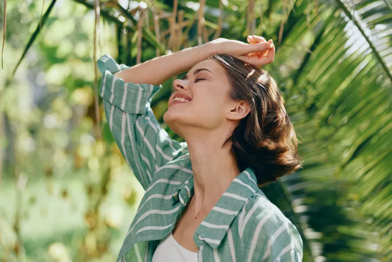 A joyful woman enjoys sunny outdoors, smiling with eyes closed as warm light highlights her relaxed pose.
